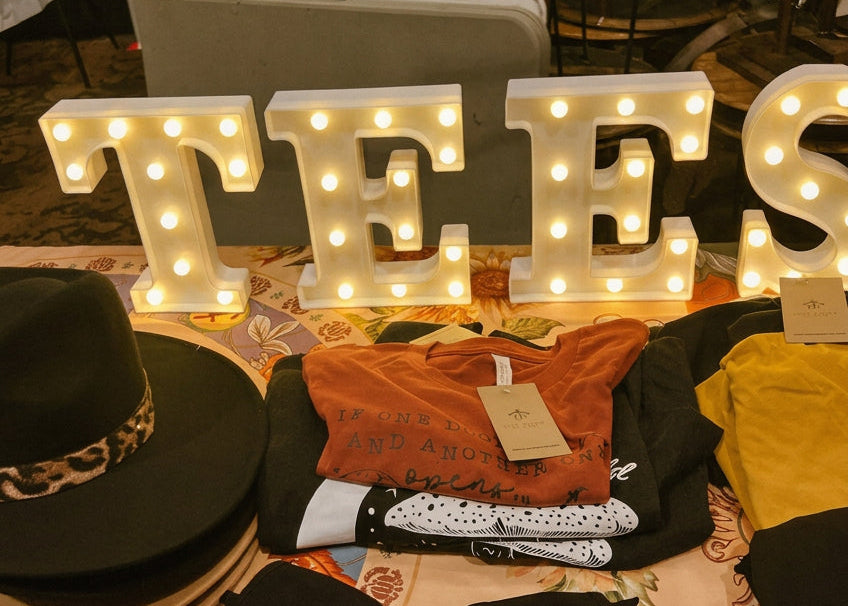 A display of folded apparel and accessories on a decorated table, featuring illuminated marquee letters spelling 'TEES', a black wide-brim hat with a leopard print band, folded orange, yellow, black, and white patterned shirts, and black pants in a backstage or event setup.